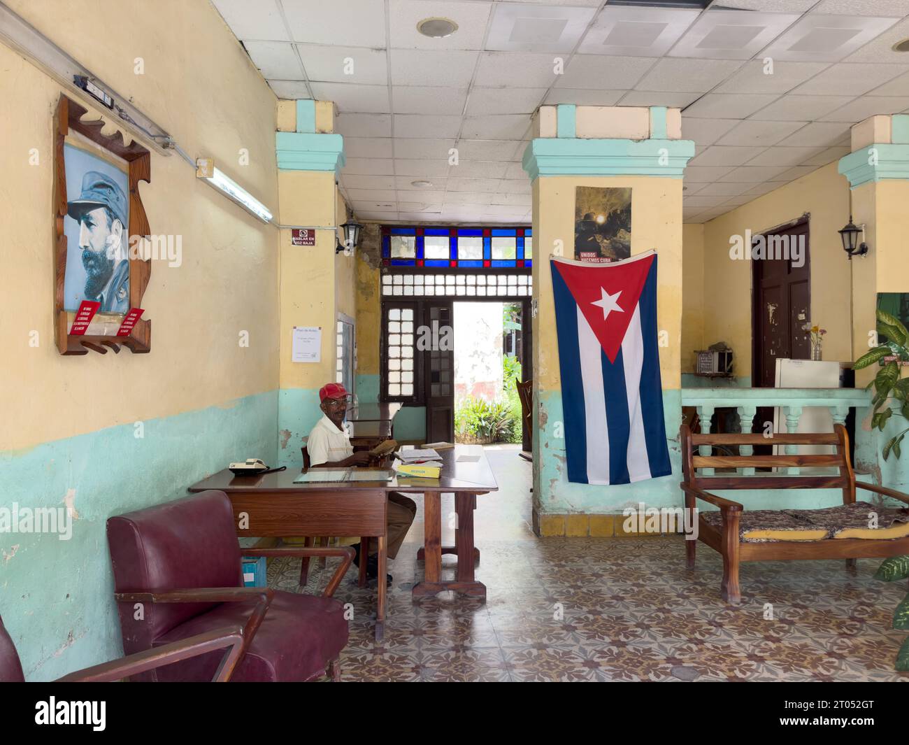 A senior Cuban man in a desk at the entrance of a government office. There is a Cuban flag and a photo of Fidel Castro decorating the hall.Santa Clara Stock Photo