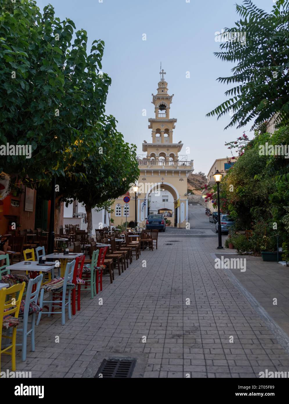 Evangelistra Church Palaiochora Chania, Crete island, Greece. Outdoors cafe on paved street, arched belfry entrance in the afternoon. Vertical Stock Photo