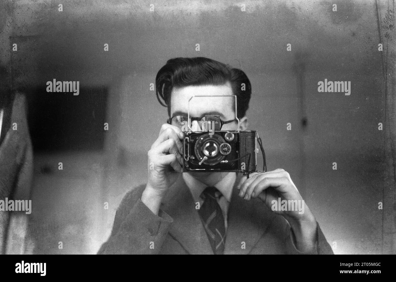 1950s, historical, a young man using a folding film camera to take a self portrait in a mirror, England, UK. Wording on the camera, IBSOR DRP, with a Anastigmat lens, suggesting that he is using a German made folding camera, which took 120 film, possibly a Rodenstock. The folding photographic camera, also known as the bellows camera, was invented by Belfast born, British architect Francis Fowkel in 1856. Stock Photo