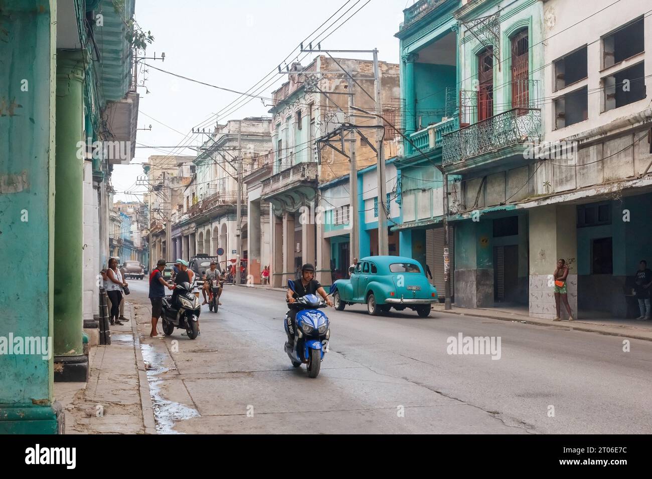 A person drives an electric bicycle on a city avenue. Other incidental people are in the street scene. Weathered buildings to both sides of the urban Stock Photo