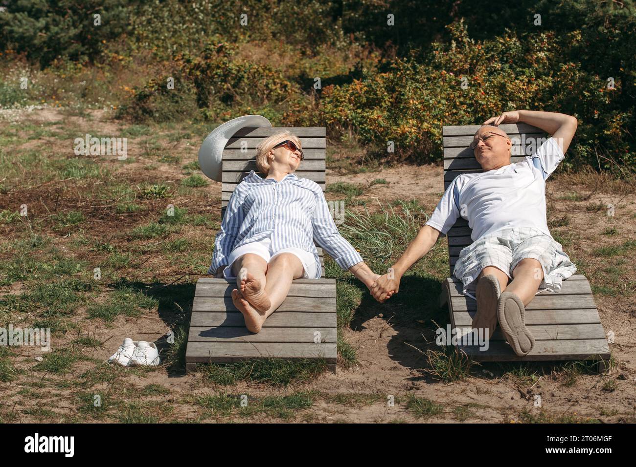 A couple of cute elderly people lie on sunbeds holding hands, basking in the sun. Stock Photo