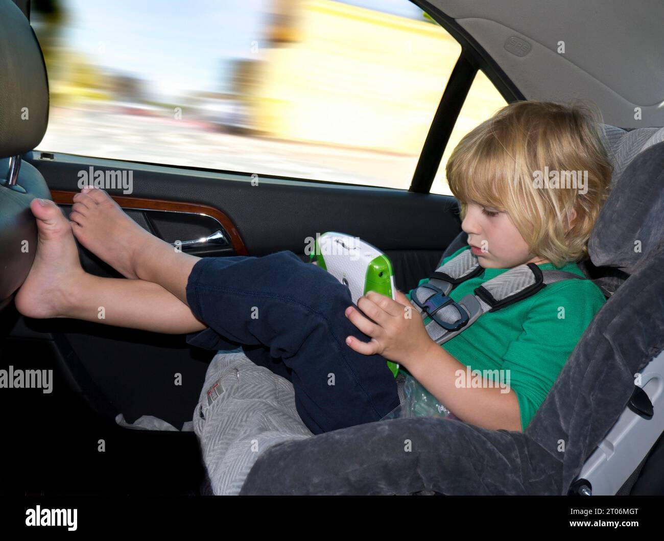 Car safety booster seat with young 5-7 years boy wearing a secure child safety belt, safely playing with his electronic gaming device in the rear of the car Stock Photo