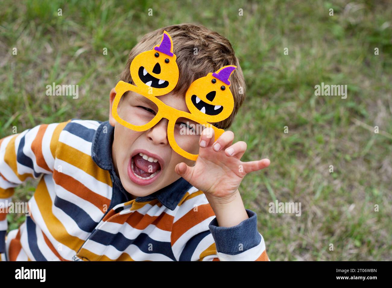 Portrait of cute boy wearing funny carnival orange glasses with pumpkins and grimacing in park. Happy Halloween. View from above. Stock Photo