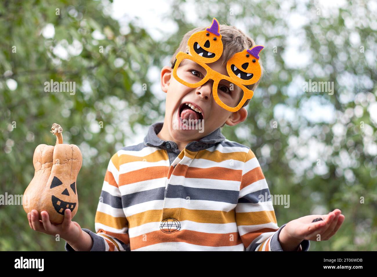 Portrait of cute boy in funny orange glasses holding a little jack lantern in his hands and grimacing in park. Happy Halloween. Stock Photo