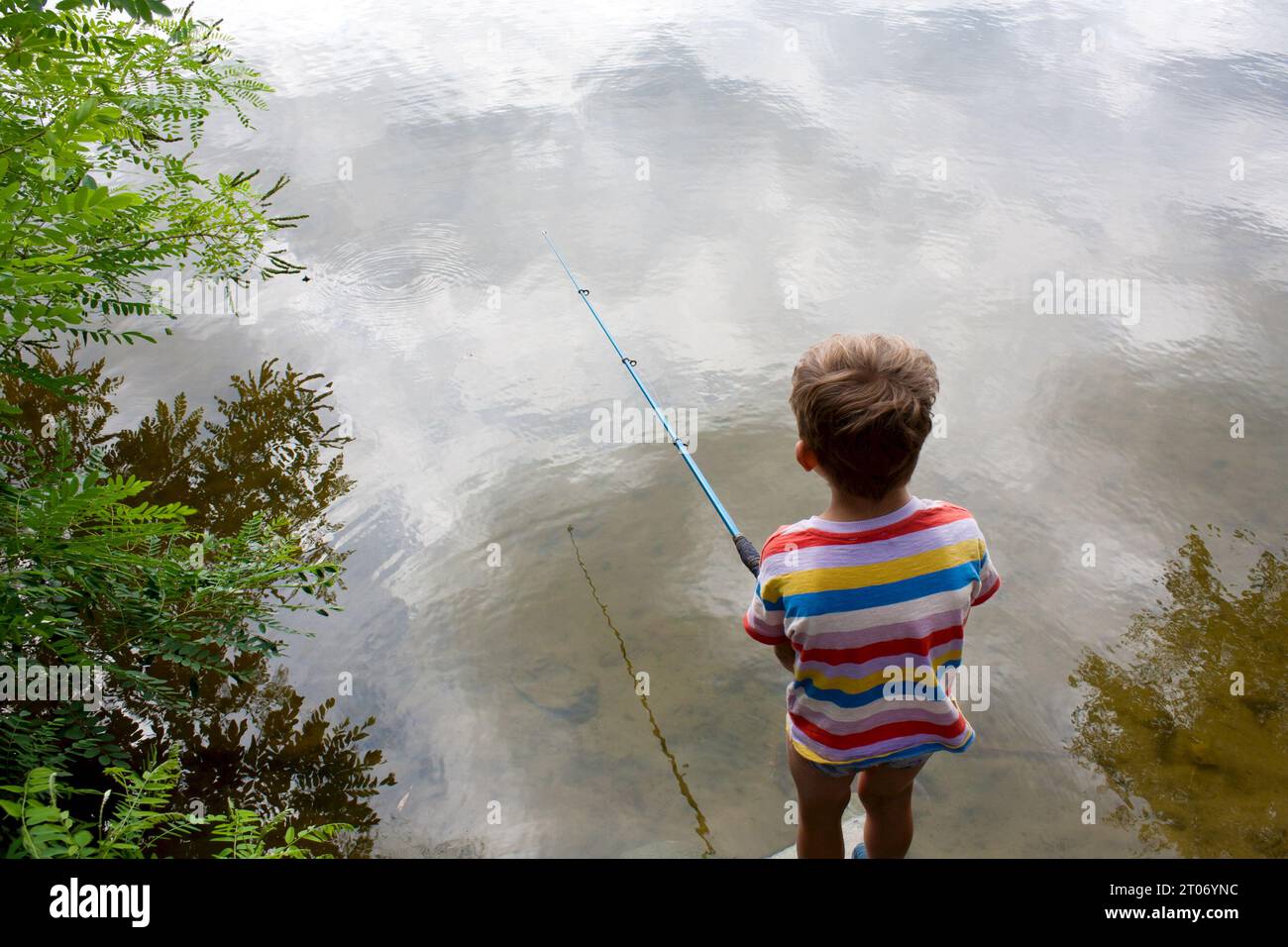 preschool child is fishing in river on cloudy summer day. boy threw fishing rod, stands with his back to camera, clouds are reflected in river. Nature Stock Photo