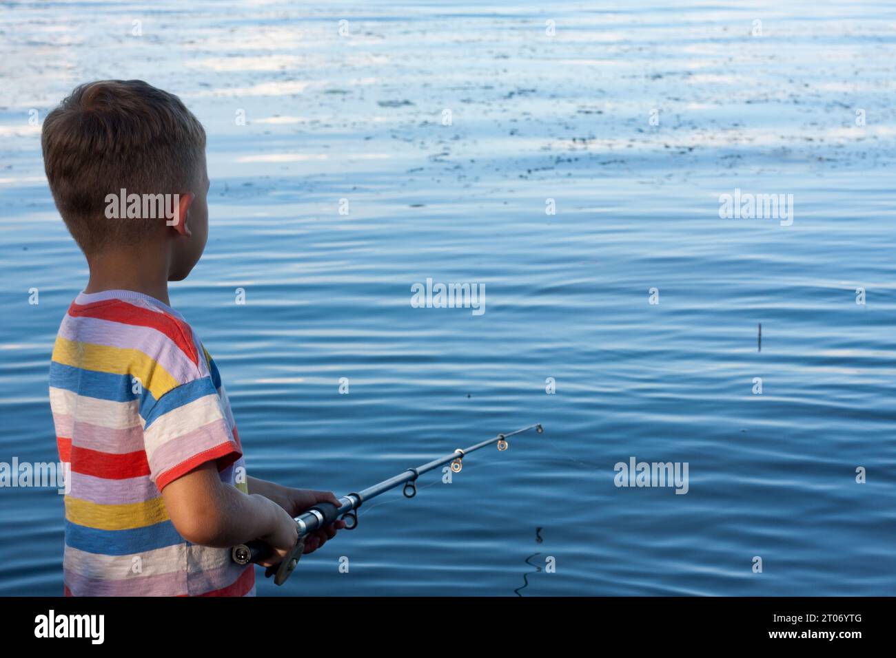 preschool child is fishing in river on summer day. boy has thrown bait and is waiting for bite, standing with his back to camera. Amateur fishing in o Stock Photo
