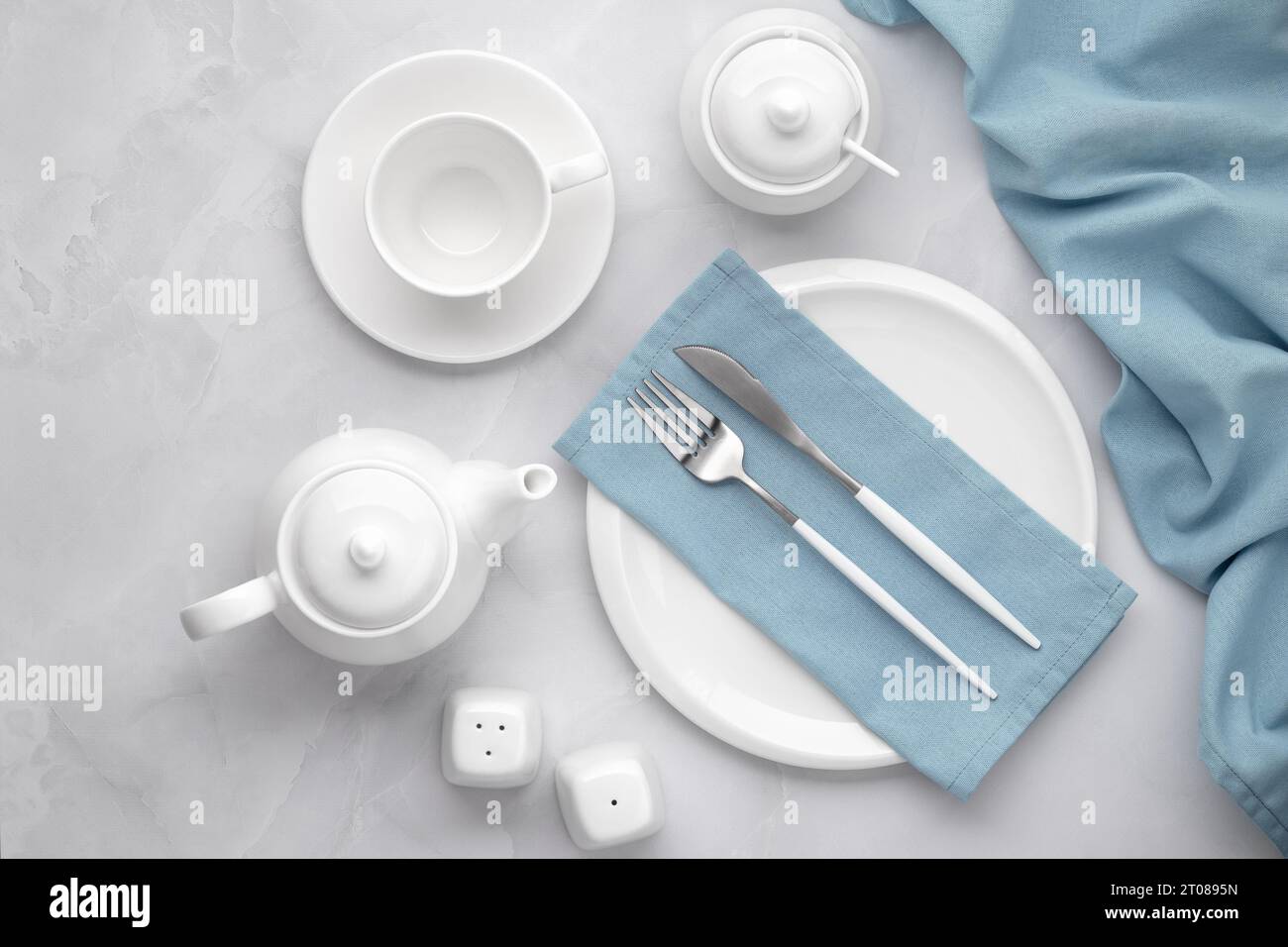 White ceramic dishes and silver cutlery. Knife, fork and blue napkin on grey background. Tea drinking in the morning. Table setting, flat lay, top vie Stock Photo