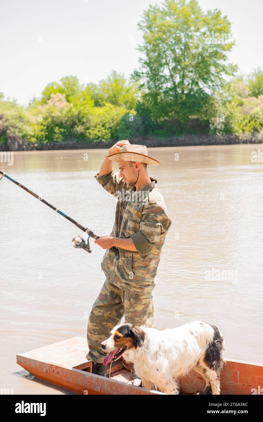 Pensive male fisher in fatigue clothes adjusting hat, while fishing with dog  on vacation Stock Photo - Alamy