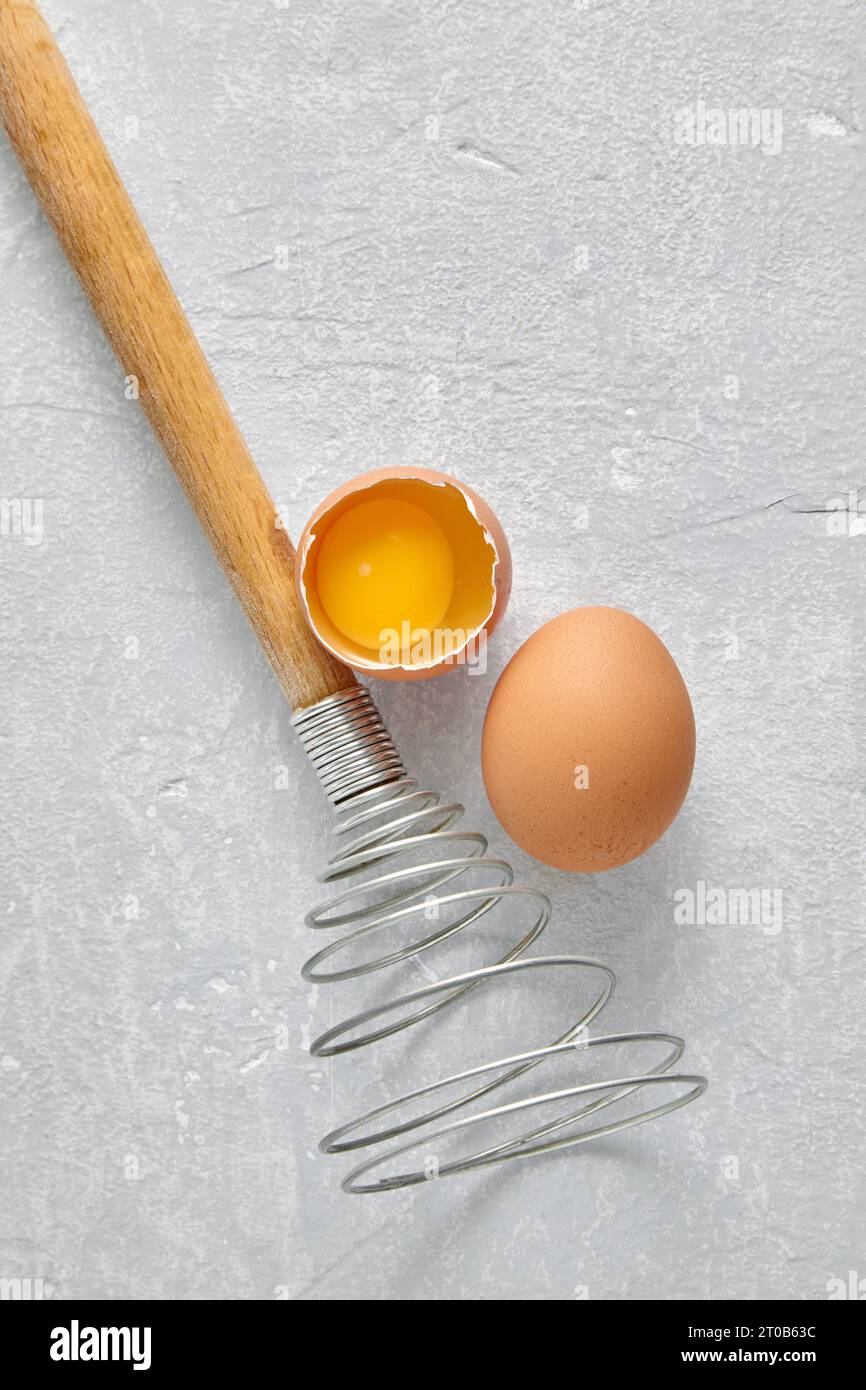 Spiral whisk and two raw beige eggs with yolk on a light concrete table. Top view with copy space Stock Photo