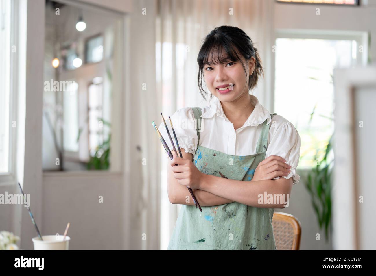 A beautiful and creative young Asian female artist in an apron smiles at the camera, stands with her arms crossed, and holds paint brushes in her hand Stock Photo