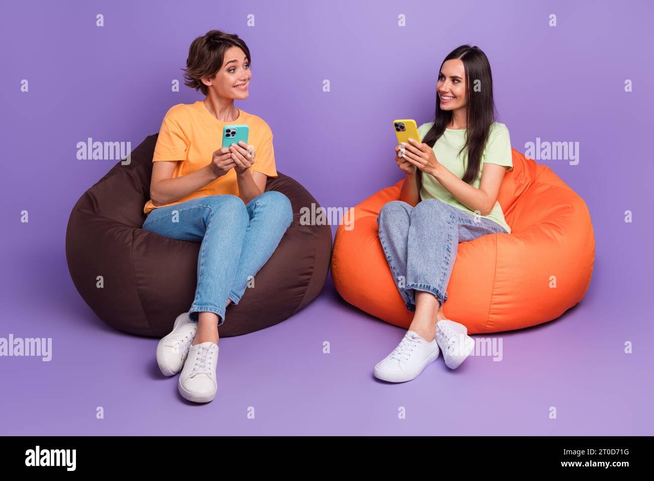 Full body length photo of sit beanbag friendship two girls using their iphones together communicating isolated on purple color background Stock Photo