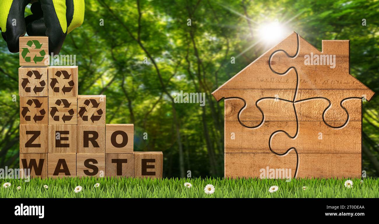 Gloved hand arranging wood blocks with text Zero Waste and Recycling Symbols on a green meadow with a small wooden house made of jigsaw puzzle pieces. Stock Photo