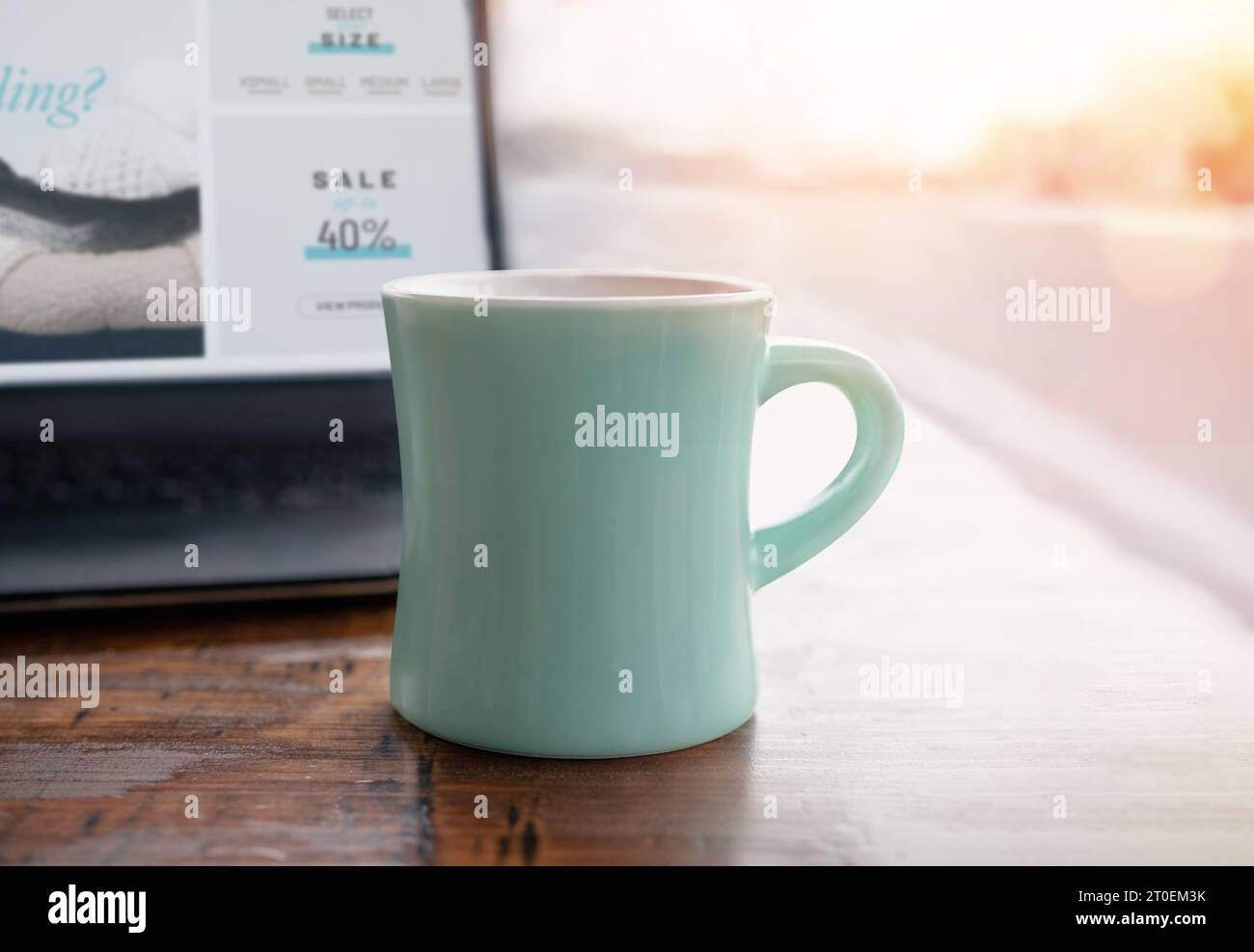 Coffee mug in front of laptop in coffee shop with defocused street scenery. White ceramic cup on wood table. Fake shopping website screen. Work from c Stock Photo
