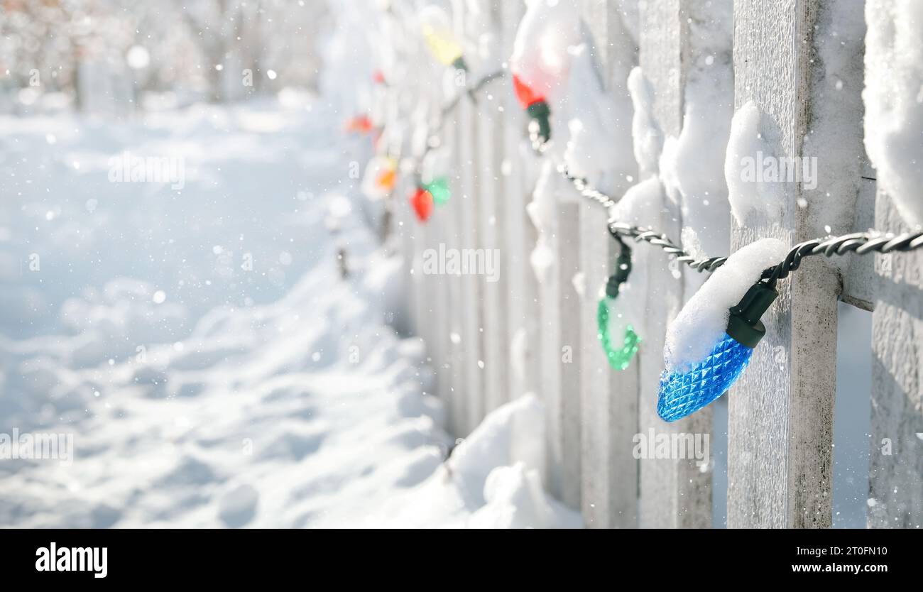 Outdoor Christmas light decoration on fence with snow flakes falling. Idyllic snowy Xmas holiday background with wooden fence and snowed in sidewalk. Stock Photo