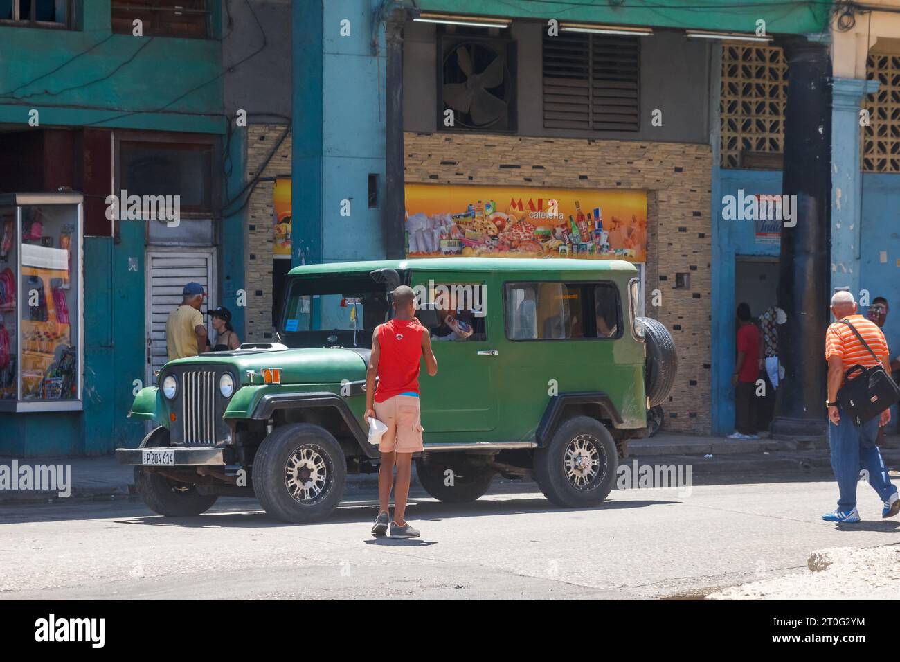 Old vintage jeep style motor vehicle driving on a city street. Cuban people are out and about the urban scene. Stock Photo
