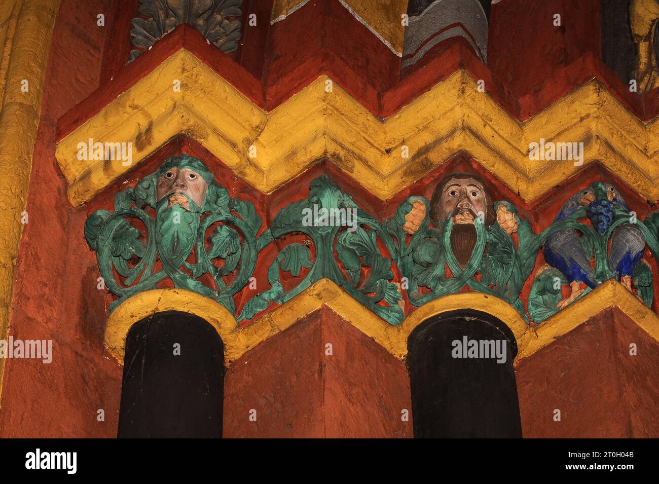 Sculpted capitals on left side of main portal of medieval Cathedral of Saint George in Limburg-an-der-Lahn, Hesse, Germany.  A bird pecks at a bunch of blue grapes (right) and faces stare out from green foliage, against a backdrop of vivid orange-red and yellow.  This Late Romanesque / Early Gothic cathedral, built in the late 1100s / early 1200s AD, was given back its original exuberant and colourful appearance through restoration work in the 1960s and ‘70s, with colours determined by traces of original paint. Stock Photo