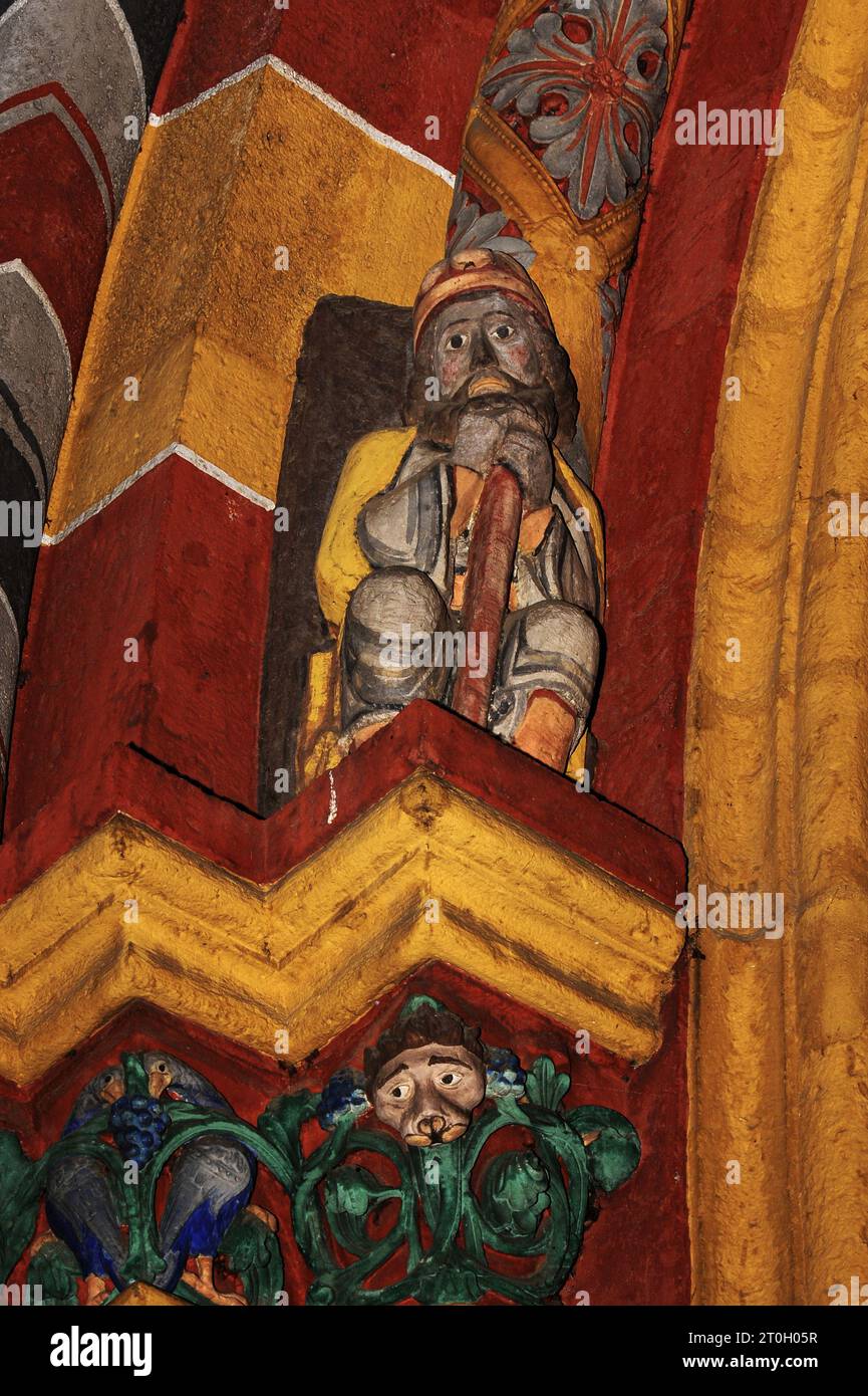 Figure of Saint Nicholas sits above sculpted capitals on left side of main portal of medieval Cathedral of Saint George in Limburg-an-der-Lahn, Hesse, Germany.  Depicted on the capitals are a bird pecking at a bunch of blue grapes (left) and a human face surrounded by green foliage, against a background of vivid orange-red and yellow.  This Late Romanesque / Early Gothic cathedral, built in the late 1100s / early 1200s AD, was given back its original exuberant and colourful appearance through restoration work in the 1960s and ‘70s, with colours determined by traces of original paint. Stock Photo