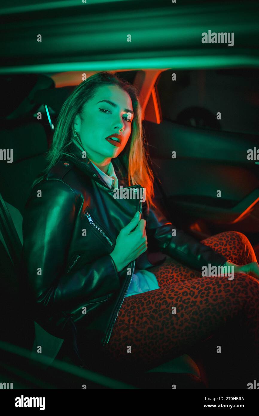 Urban photography with green and red neons in a car. Young blonde Caucasian model pose in a black leather jacket Stock Photo