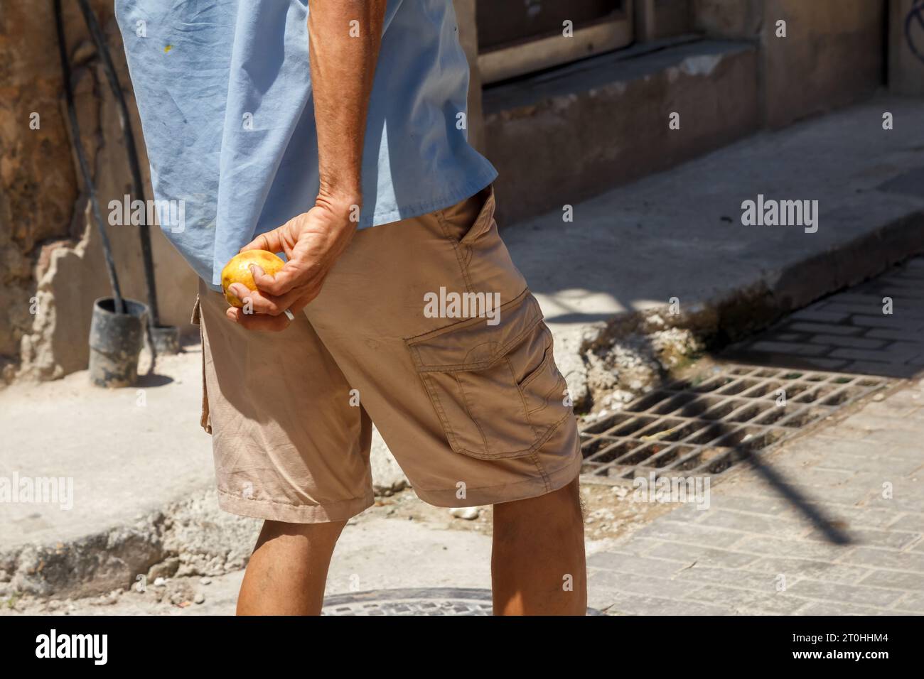 A Cuban man holds a ripe guava fruit in his hand as he walks on a city street in Old Havana. The sidewalk is damaged in the corner. Stock Photo