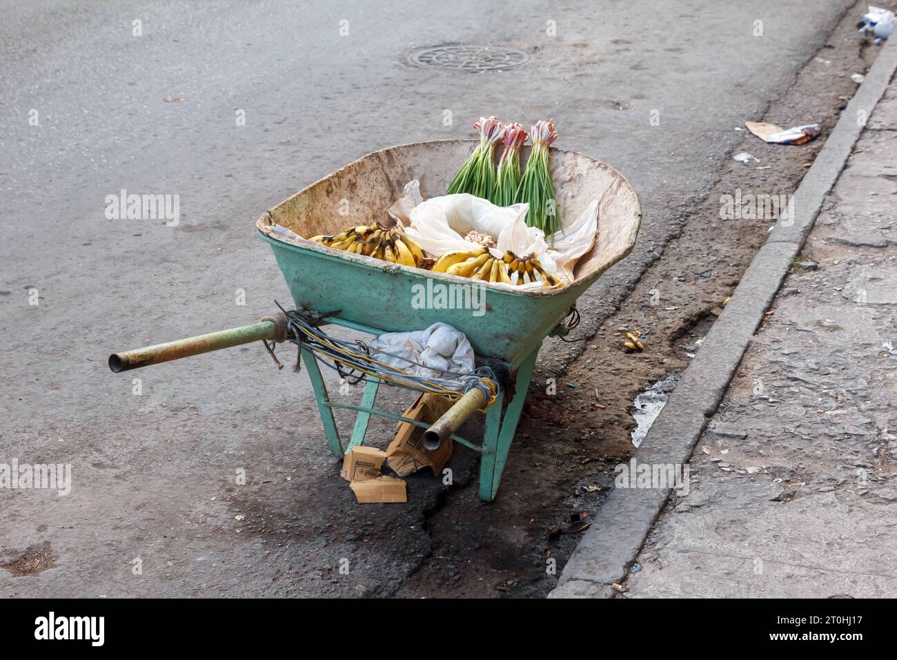 A wheelbarrow with fruit and spices for sale. The object is stationary by the dirty curb of a city street. The sidewalk is damaged. Stock Photo