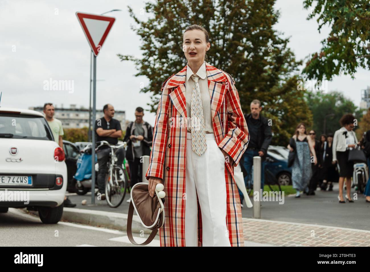 A guest wearing a checkered trench coat, white pants, a tie and brown bag, seen outside PRADA show during Milan Fashion Week Womenswear Spring/Summer Stock Photo