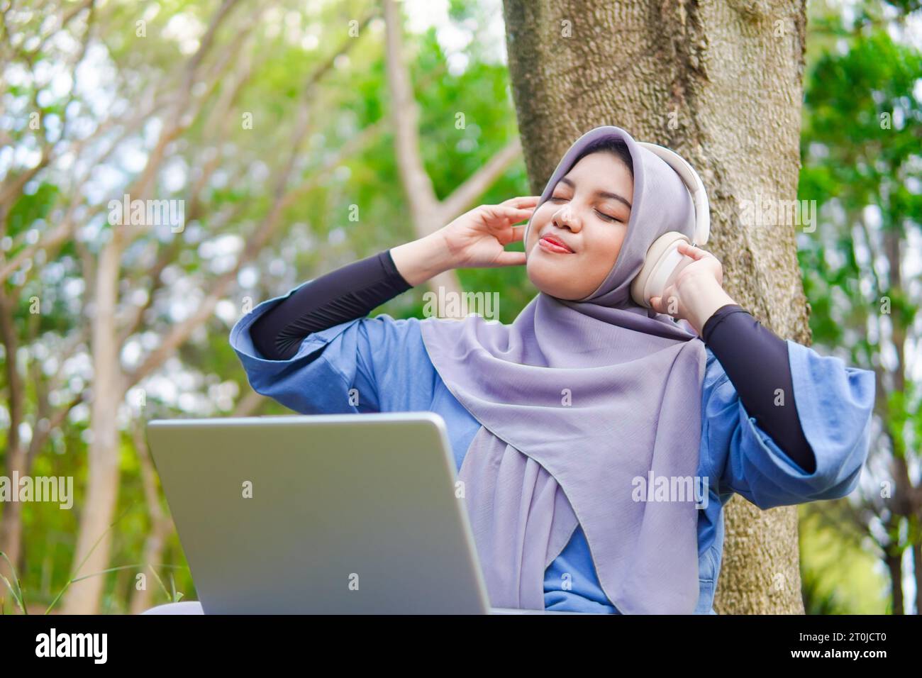 muslim woman with laptop and wearing headphone. listening to music in the park. under the tree Stock Photo