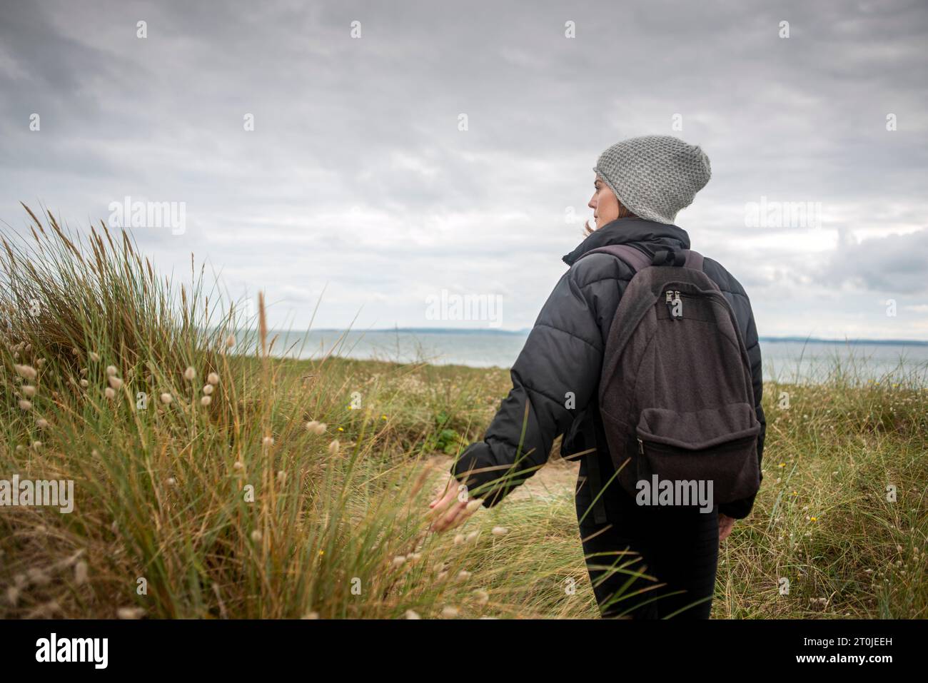 woman walking through sand dunes by the sea wearing a wooly hat and a jacket, winter healthy lifestyle. Stock Photo