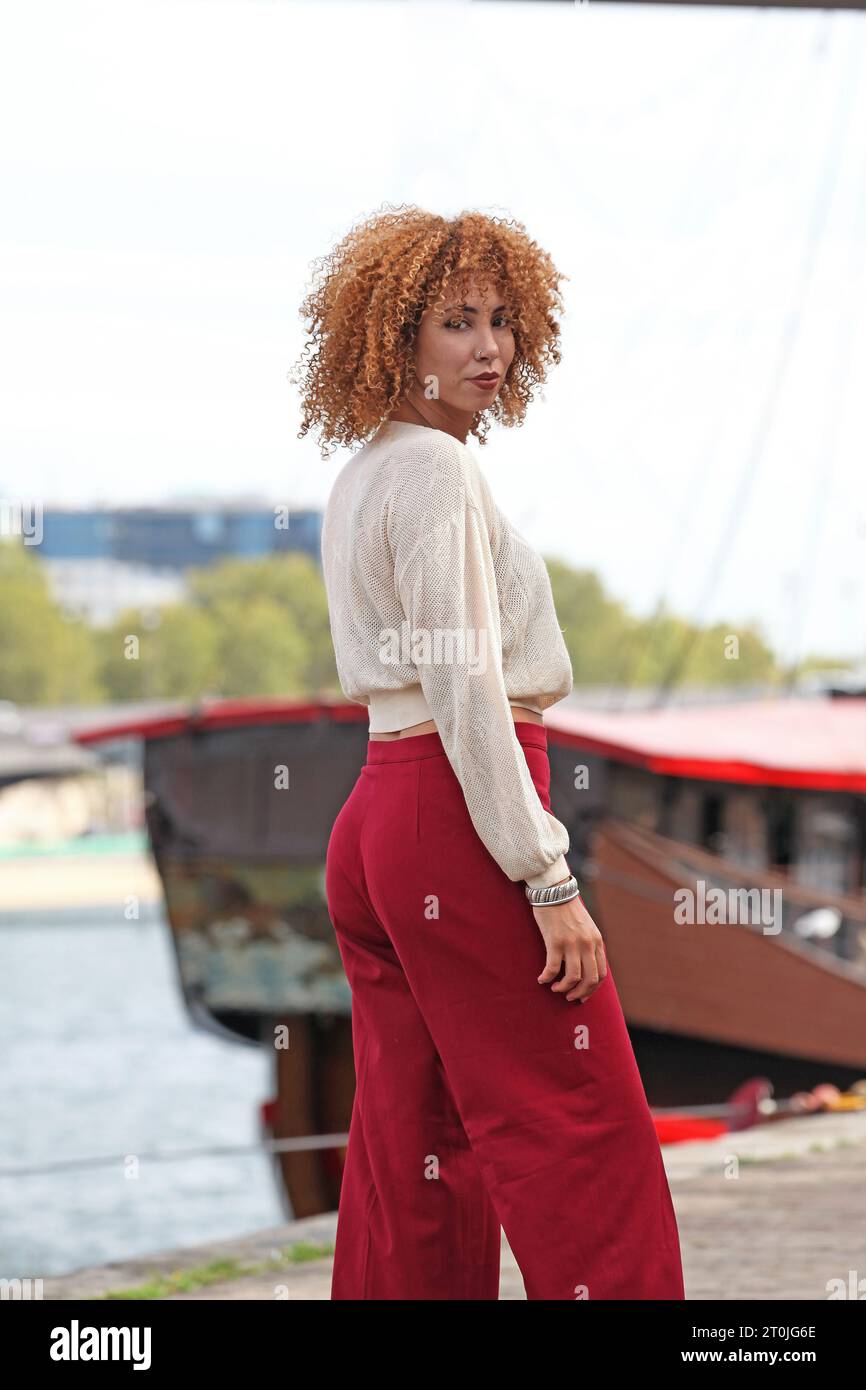woman with curly hair and red pants posing on a dock near a boat Stock Photo