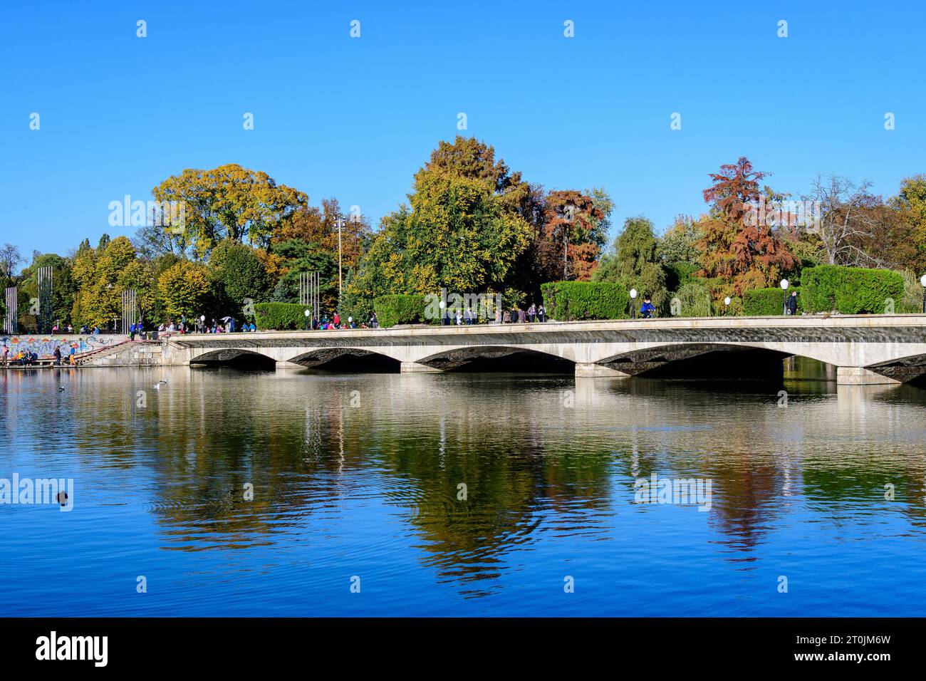 Landscape with long old grey bridge and many large green trees near the lake in Carol Park in Bucharest, Romania,  in a sunny autumn day Stock Photo