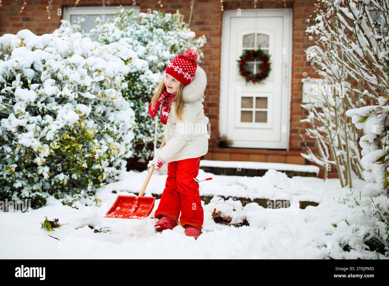 Child shoveling snow. Little girl with spade clearing driveway after winter snowstorm. Kids clear path to house door after Christmas blizzard. Stock Photo
