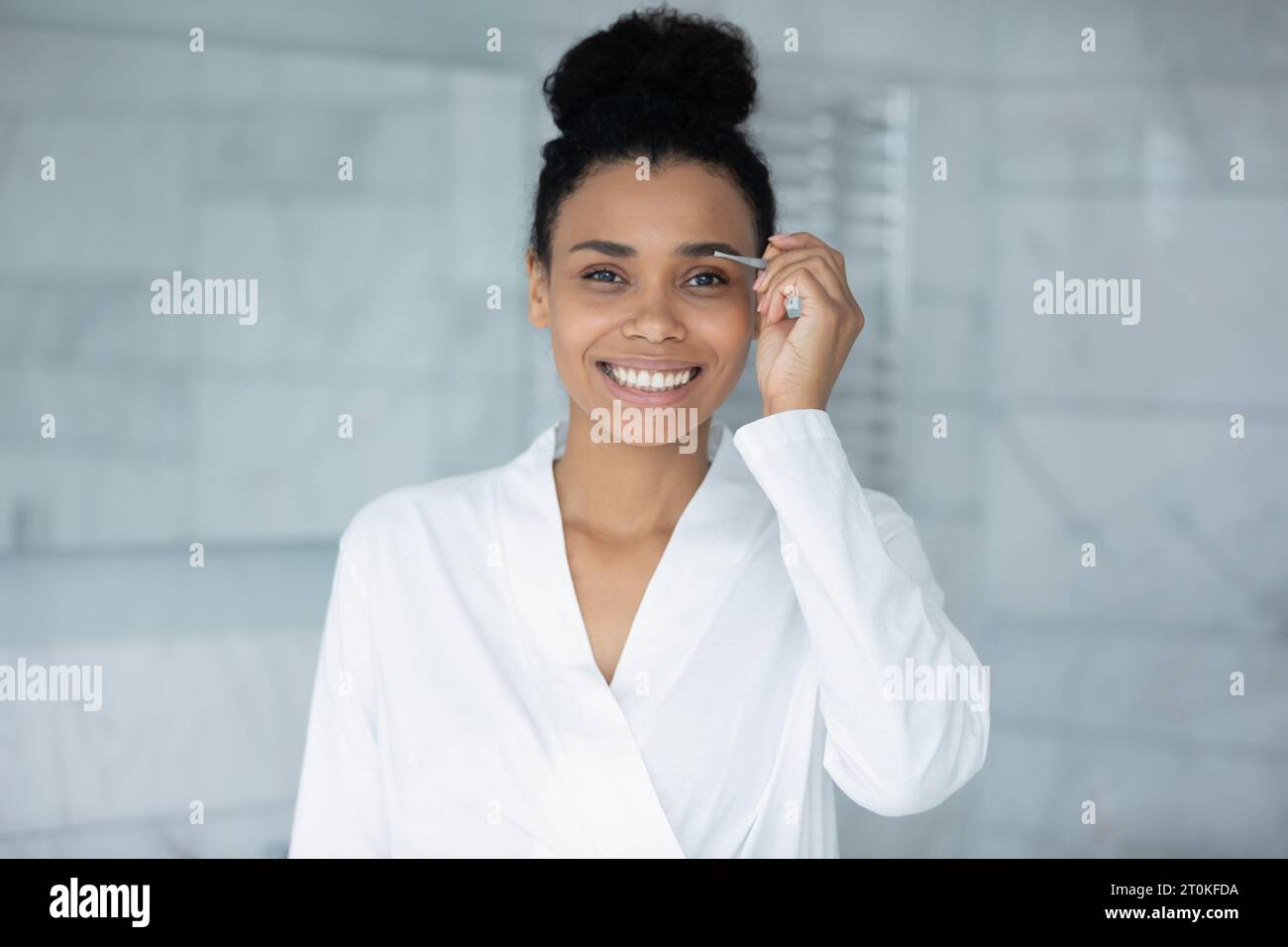 Smiling biracial woman pinch eyebrows with tweezers Stock Photo