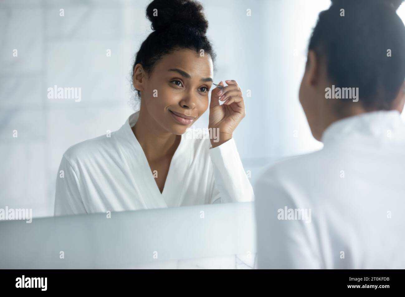 African American woman pinch eyebrows with tweezers in bathroom Stock Photo