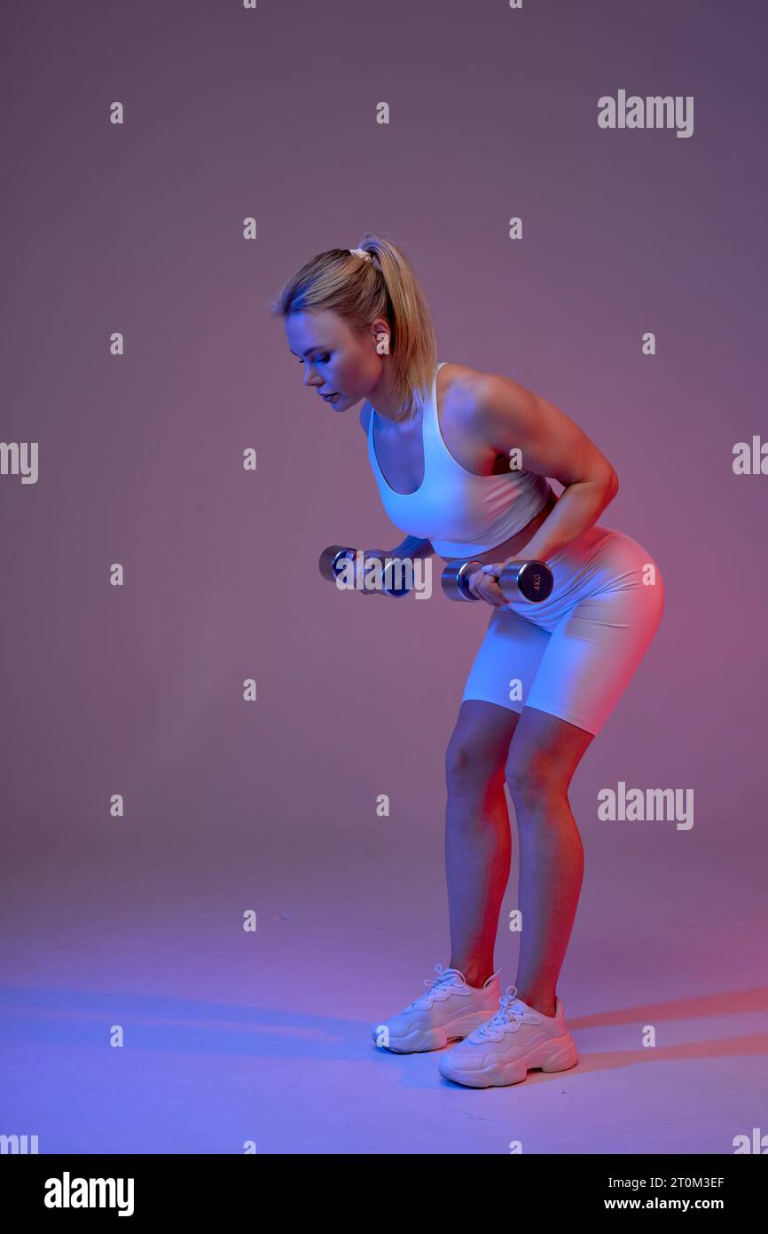 Side view of fit healthy fair-haired girl in sportswear bending and doing exercises with dumbbells in hands. isolated purple background Stock Photo