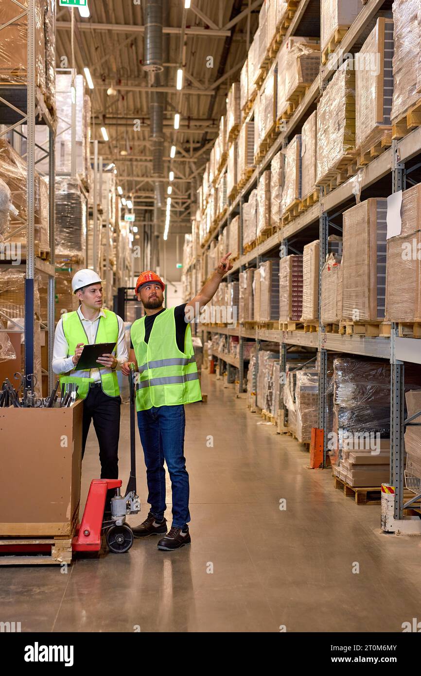 two men warehouse workers with hand pallet jack lift stand having talk, temwork. hand pallet truck and male mover. Workers in uniform with cargo cart. Stock Photo