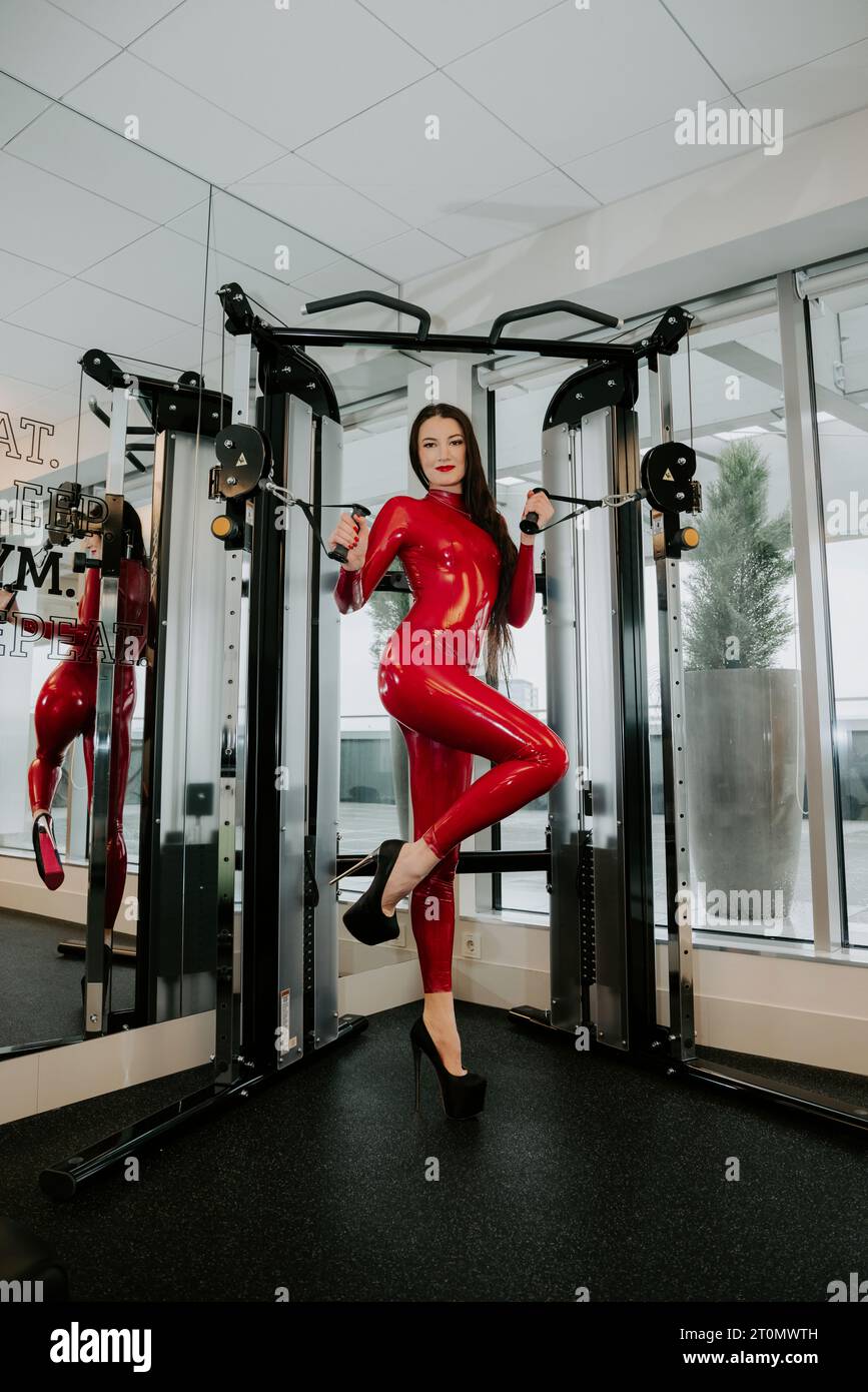 Brunette female wearing a shiny red latex catsuit and high heels is standing in fitness apparatus Stock Photo