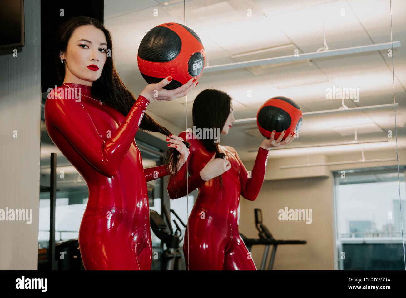 Brunette female wearing a shiny red latex catsuit with a training ball in her hand is looking at us, standing in front of a mirror Stock Photo