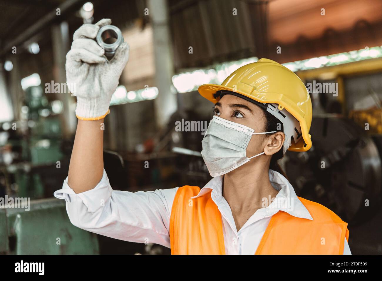Young Asian woman worker hard working with heavy metal machine lathe milling factory industry dirty workplace waring face mask for hygiene. Stock Photo