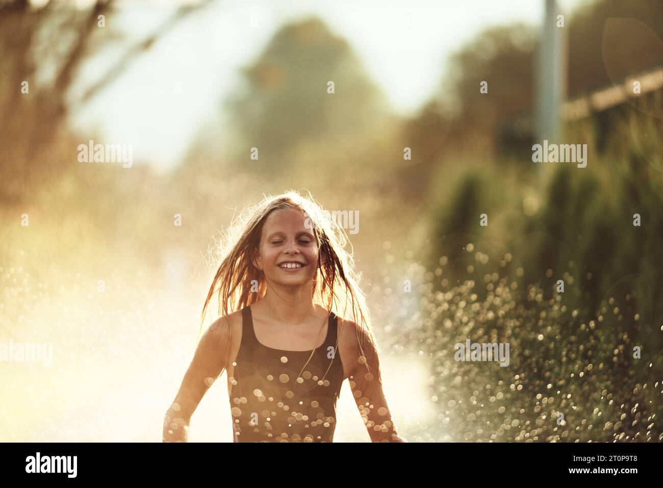 In the backyard of house, a young girl bursts with laughter and joy as she gleefully runs through a water sprinkler on a sunny summer day Stock Photo