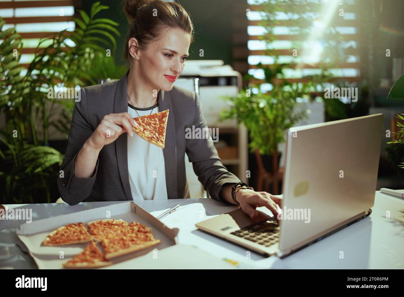 Sustainable workplace. modern small business owner woman in a grey business suit in modern green office with pizza and laptop. Stock Photo