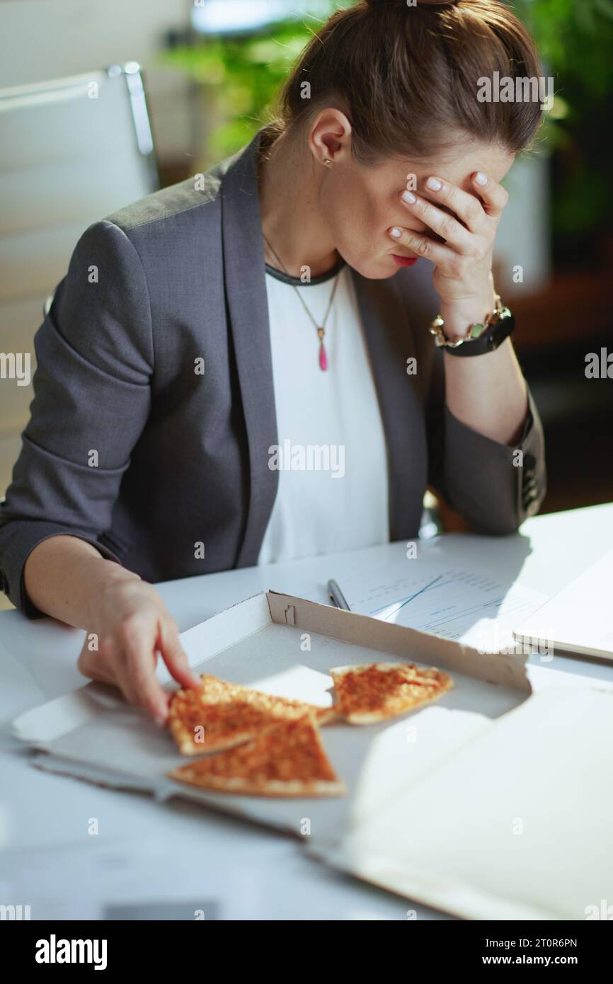 Sustainable workplace. stressed modern middle aged bookkeeper woman in a grey business suit in modern green office with pizza. Stock Photo