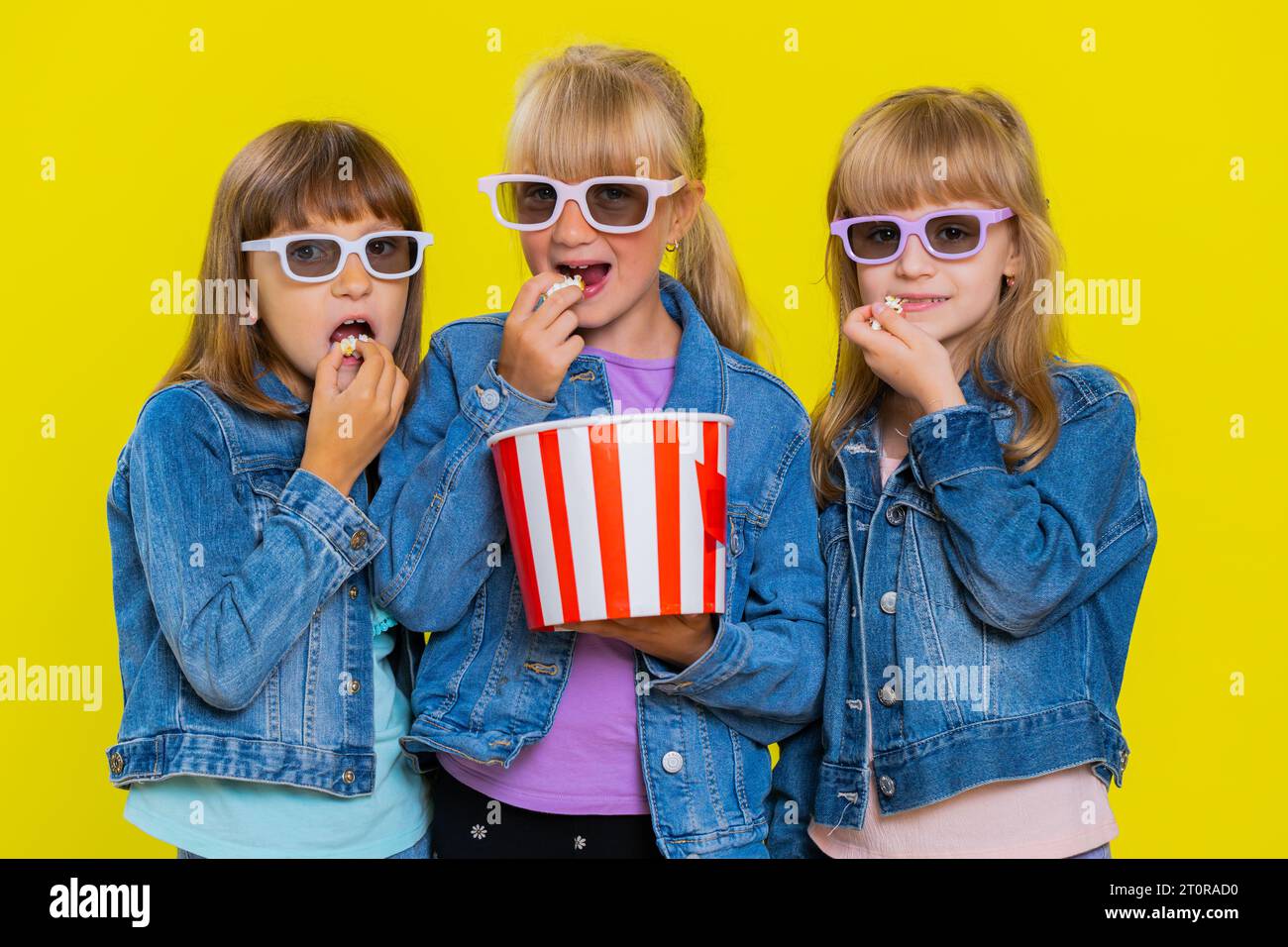 Teenage girls wearing 3D glasses eating popcorn watching interesting tv serial, sport game, film, online social media movie content. Little children sisters. Three siblings kids on yellow background Stock Photo