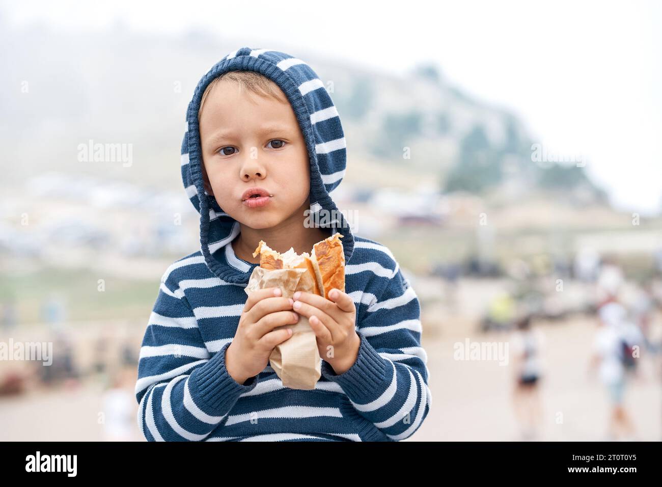 Lovely little boy holds tasty hotdog walking in mountain park. Child eating healthy snack on travel portrait. Calm kid with sandwich outdoors Stock Photo