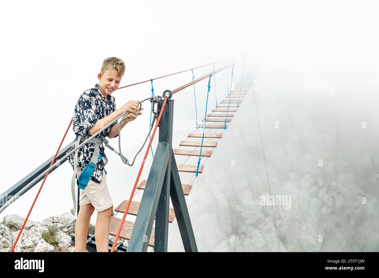 Smiling boy fastens safety belt carabine preparing to walk along rope bridge at mountain. Happy kid stands near extreme attraction in misty valley Stock Photo