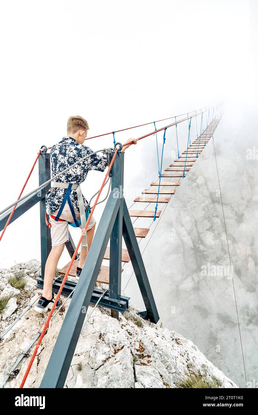 Courageous boy walks along rope bridge in mountain park on foggy day. Little tourist enjoys extreme attraction in cliffy valley. Sports activity Stock Photo