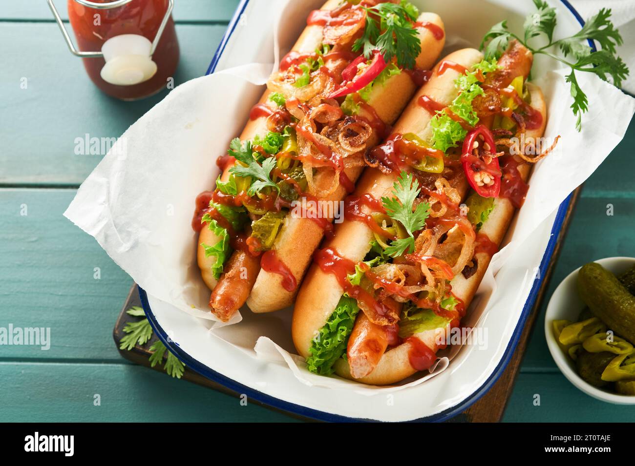 Delicious grilled hotdog with pickled  cucumbers, chili peppers, caramelized onions, ketchup, mustard in craft paper on blue old wooden background. Ho Stock Photo