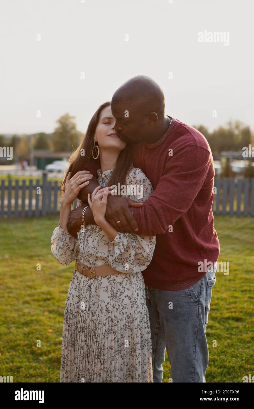 Portrait of interracial couple in love, gently hugging and kissing with eyes closed in nature park. Man embrace girlfriend from behind, standing toget Stock Photo