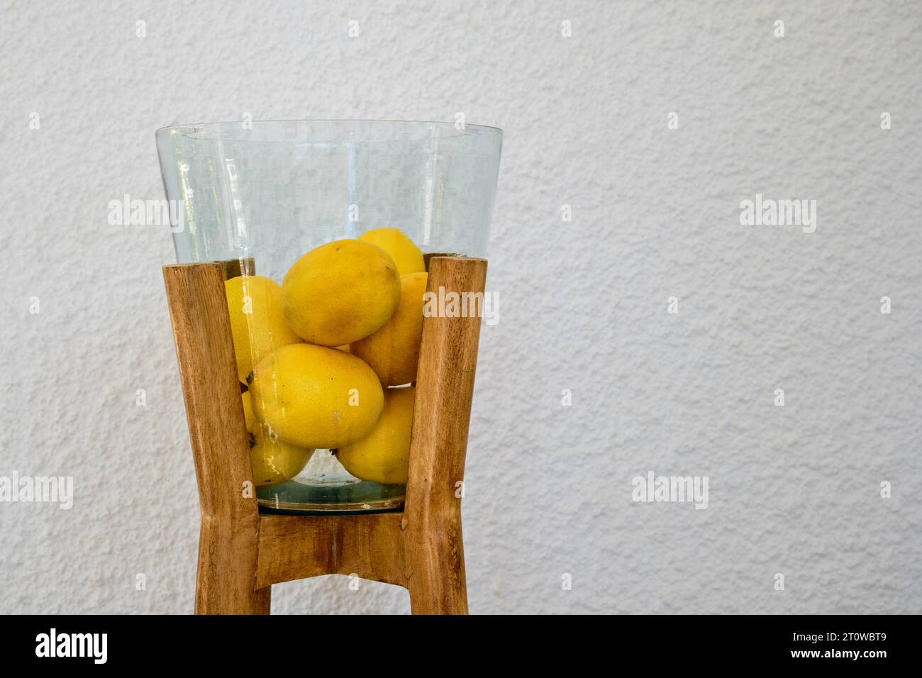 Decorative glass bowl or vase with wooden feet filled with fresh lemons against white wall background. Stock Photo