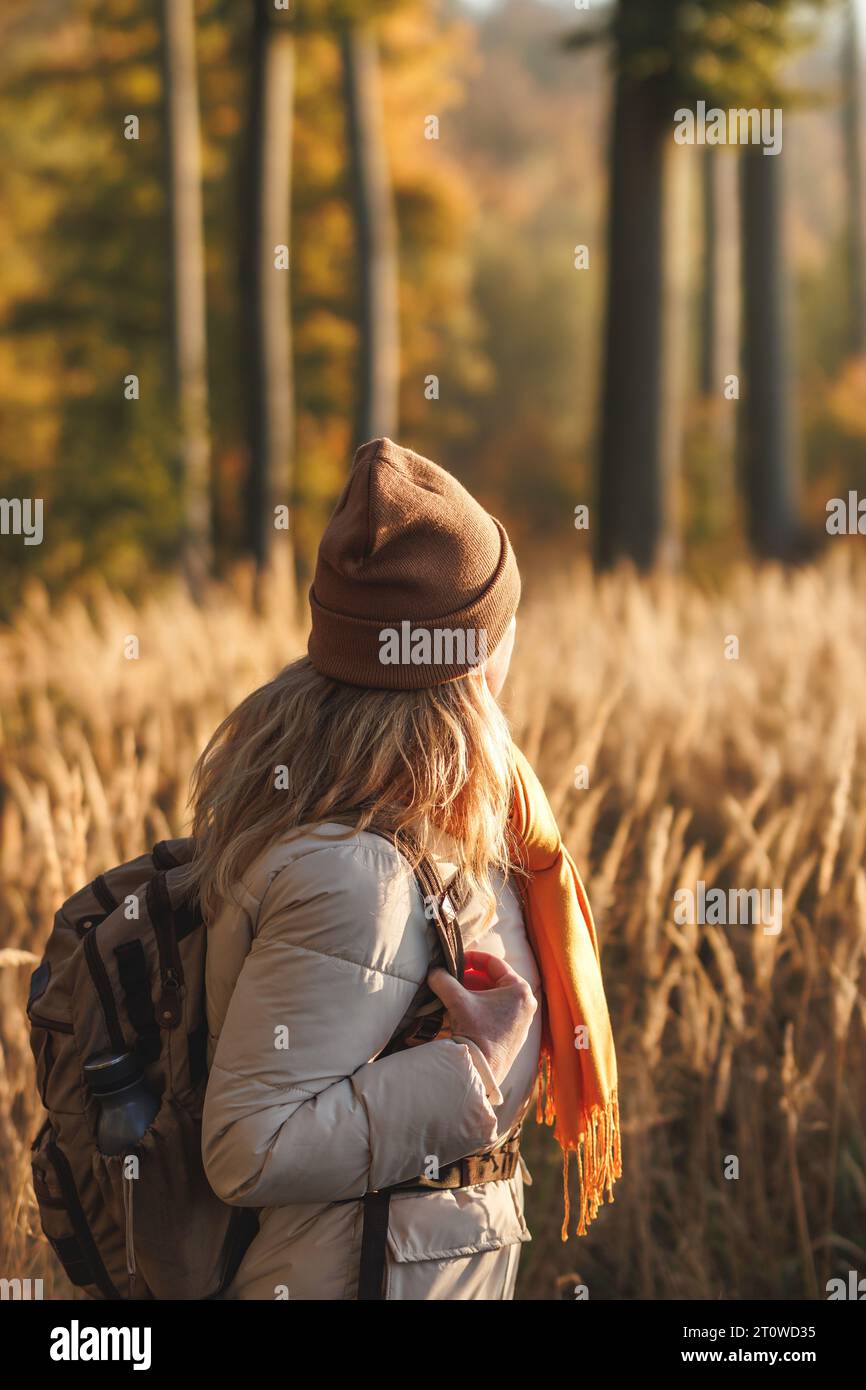 Woman with backpack, jacket, scarf and knit hat walks in golden grass. Autumn hiking in forest. Outdoor warm clothing Stock Photo