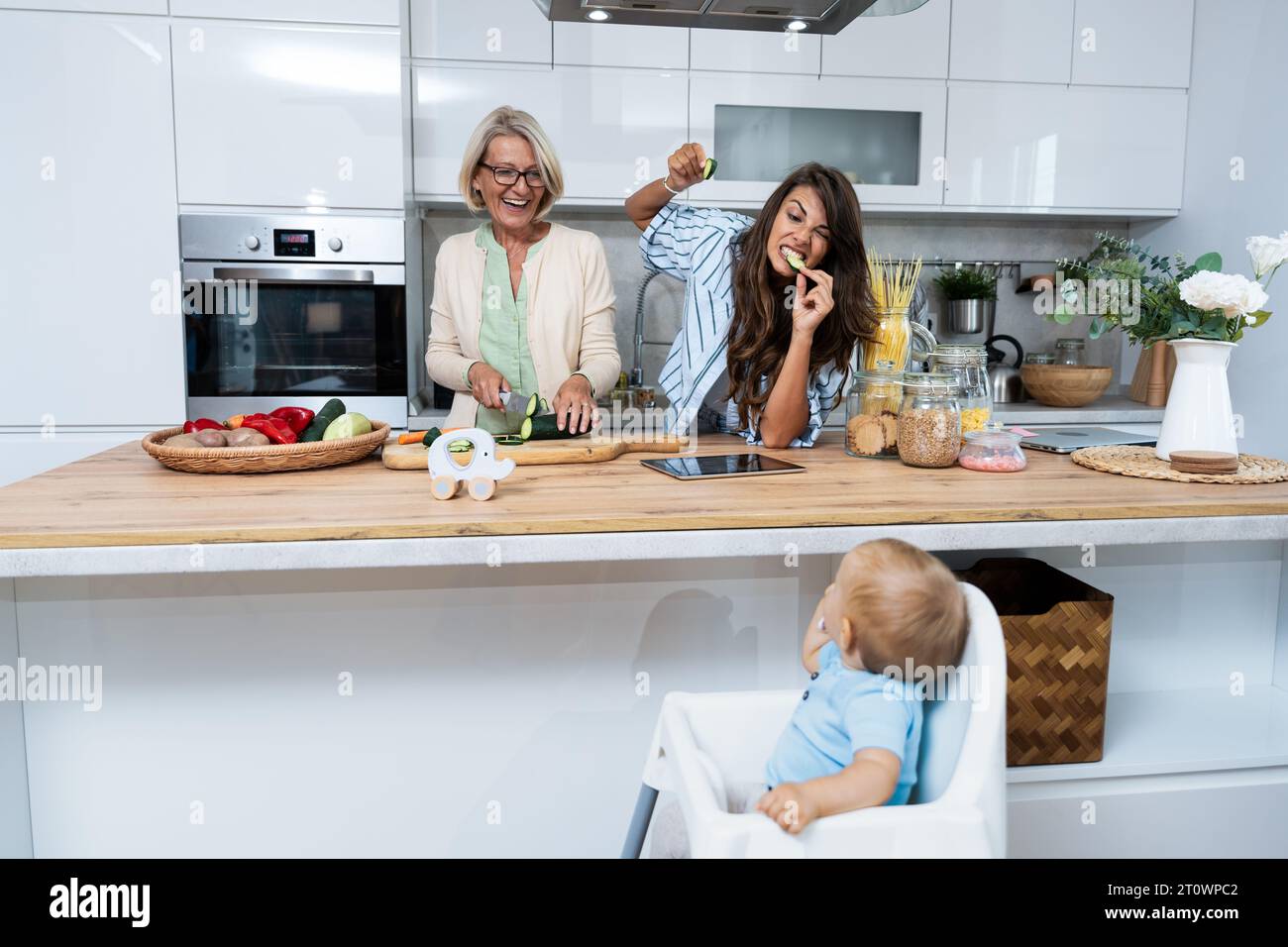Grandmother mother and baby granddaughter cooking together in kitchen in apartment while mom making a funny faces for child and granny cutting vegetab Stock Photo