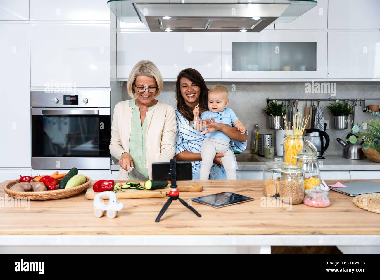 Grandmother mother and baby granddaughter cooking together in kitchen in apartment while mom making a funny faces for child and granny cutting vegetab Stock Photo